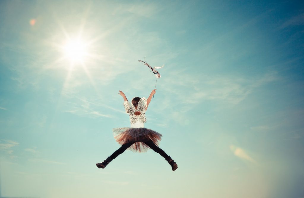 Little girl in jeans, boots, tutu, fairy wings, and wand, jumping high into the open sky.
